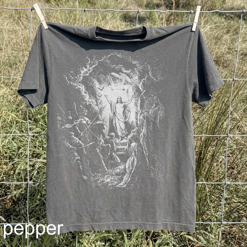 May include: Dark gray t-shirt with a white graphic depicting a religious scene. The graphic shows a figure with arms raised, surrounded by other figures and light. The word "pepper" is printed at the bottom.