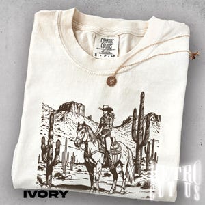 May include: Ivory-colored t-shirt with a graphic of a cowgirl on a horse in a desert landscape with cacti. The shirt has the text "IVORY" at the bottom and a gold necklace. The shirt is made by Comfort Colors.