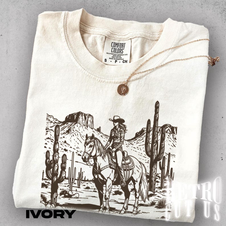 May include: Ivory-colored t-shirt with a graphic of a cowgirl on a horse in a desert landscape with cacti. The shirt has the text "IVORY" at the bottom and a gold necklace. The shirt is made by Comfort Colors.