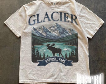 May include: Ivory-colored t-shirt featuring a scenic design with the word "GLACIER" above a mountain range, lake, and moose silhouette. The words "NATIONAL PARK" are on a banner below the moose. The shirt has short sleeves.
