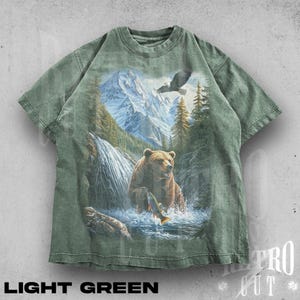 Puede incluir: Camiseta verde claro con una escena de la naturaleza: un oso pescando en un río, montañas y un águila. El texto "LIGHT GREEN" está impreso en la parte inferior.