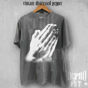 May include: A gray t-shirt with a distressed print of praying hands. The shirt has a vintage look and the text "vintage distressed pepper" is printed at the top. The shirt is hanging on a wooden hanger.