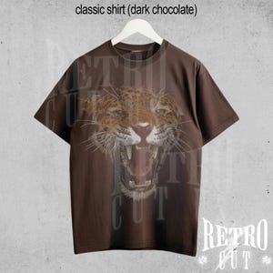 May include: A brown t-shirt featuring a graphic of a snarling jaguar on the front. The shirt has a crew neck and short sleeves. The text "Retro Cut" is printed on the bottom right corner of the shirt.