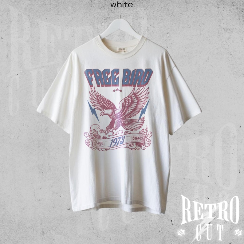 May include: A white t-shirt with a vintage-style graphic of a red eagle with blue wings and the text "FREE BIRD 1973".