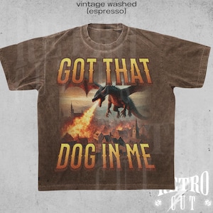 May include: A brown t-shirt with a vintage washed look features a graphic of a dragon breathing fire over a medieval town. The text "Got That Dog In Me" is printed in a bold font below the image.