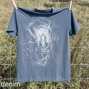 May include: Denim blue t-shirt with a white graphic print of a religious scene. The image depicts a figure with outstretched arms, surrounded by other figures, with the word "denim" printed at the bottom. The shirt is hanging on a clothesline.
