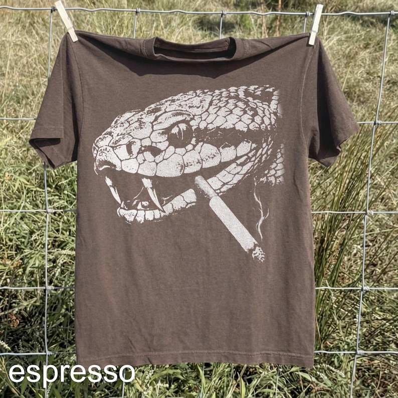 May include: Espresso-colored t-shirt featuring a detailed white graphic of a snake's head with a cigarette in its mouth. The shirt is hanging on a clothesline. The word "espresso" is printed at the bottom.
