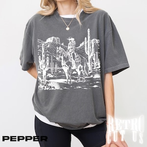 May include: Dark gray t-shirt featuring a white graphic of a person riding a horse through a desert landscape with cacti and mountains. The word "PEPPER" is printed in black at the bottom left.