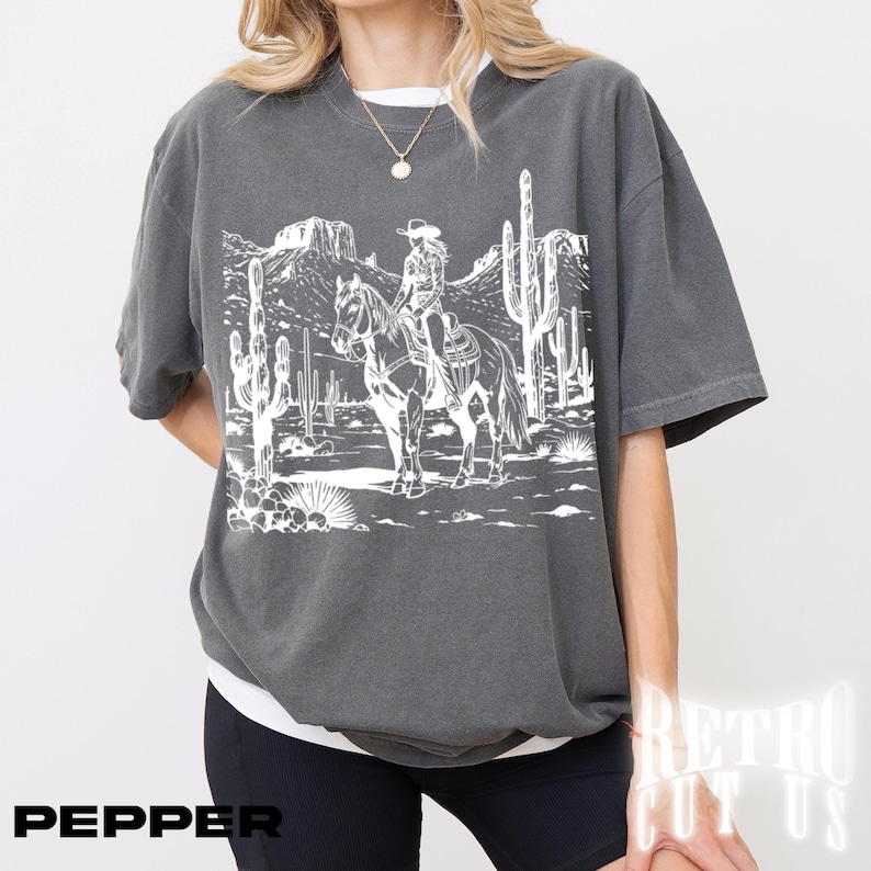 May include: Dark gray t-shirt featuring a white graphic of a person riding a horse through a desert landscape with cacti and mountains. The word "PEPPER" is printed in black at the bottom left.