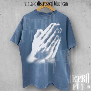 May include: A vintage distressed blue jean t-shirt with a white graphic of praying hands. The shirt is a faded blue color and has short sleeves. The text "vintage distressed blue jean" is at the top.