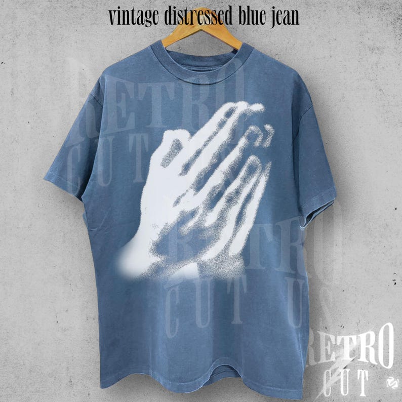 May include: A vintage distressed blue jean t-shirt with a white graphic of praying hands. The shirt is a faded blue color and has short sleeves. The text "vintage distressed blue jean" is at the top.