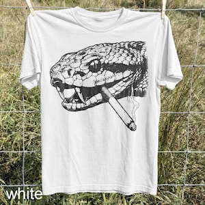 May include: White t-shirt featuring a detailed black and white illustration of a snake's head with a lit cigarette in its mouth. The shirt is hanging on a clothesline. The word "white" is printed at the bottom.