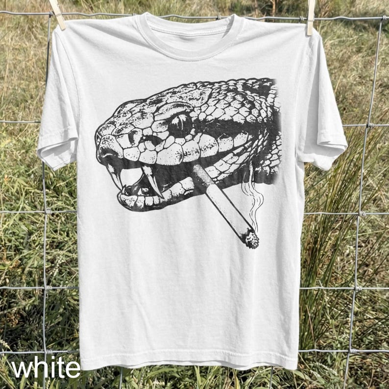 May include: White t-shirt featuring a detailed black and white illustration of a snake's head with a lit cigarette in its mouth. The shirt is hanging on a clothesline. The word "white" is printed at the bottom.