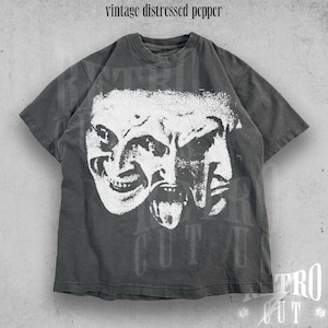May include: A dark grey t-shirt with a distressed look featuring a white graphic of two distorted faces with open mouths and tongues sticking out. The text "Retro Cut" is visible in the bottom right corner.