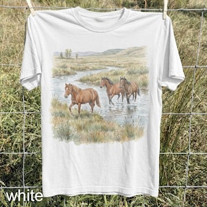 May include: White t-shirt featuring a watercolor painting of three brown horses in a stream. The horses are wading through the water, with a landscape of grassy banks and distant hills in the background. The word "white" is printed at the bottom.