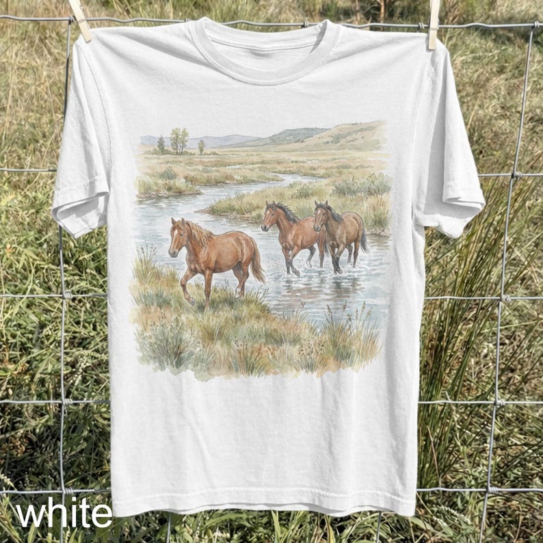 May include: White t-shirt featuring a watercolor painting of three brown horses in a stream. The horses are wading through the water, with a landscape of grassy banks and distant hills in the background. The word "white" is printed at the bottom.