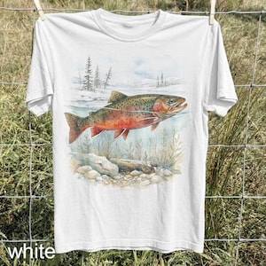 K&ouml;nnte beinhalten: Wei&szlig;es T-Shirt mit einer Aquarellillustration eines Fisches in einem Fluss. Der Fisch hat rote und braune Markierungen. Der Hintergrund zeigt eine verschneite Landschaft und Wasserpflanzen. Das Wort "white" ist unten auf dem T-Shirt aufgedruckt.