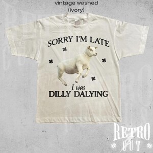 May include: A white t-shirt with a vintage washed look. The shirt features a black graphic of a sheep with the text "SORRY I'M LATE I was DILLY DALYING".