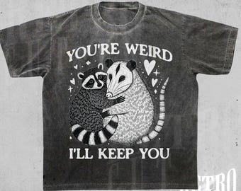 May include: A black t-shirt with a vintage washed look features a graphic of a raccoon and a possum hugging with the text "You're Weird I'll Keep You".