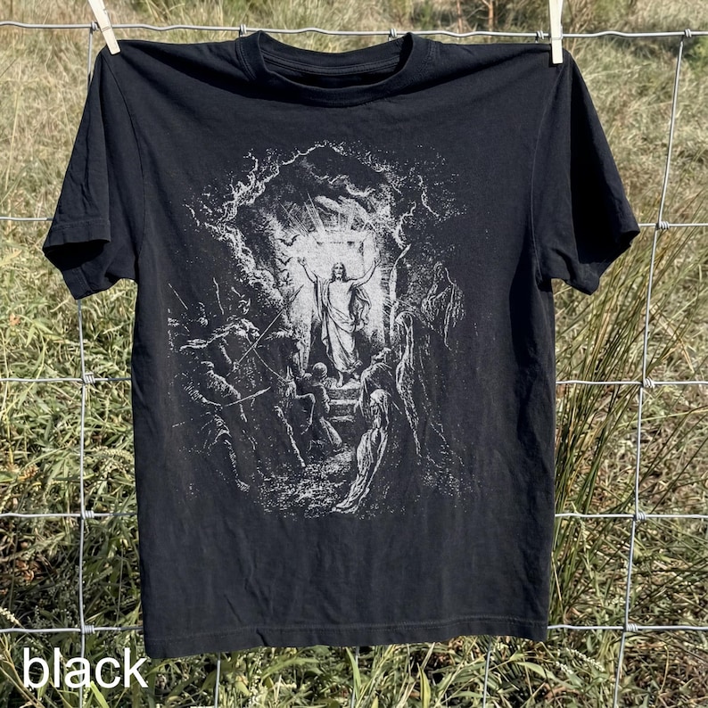 May include: Black t-shirt featuring a detailed, grayscale illustration of a religious scene. The graphic depicts a central figure with outstretched arms, surrounded by other figures and set within a cave-like structure. The word "black" is printed at the bottom.