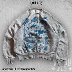May include: A gray sweatshirt with a blue print of various sea creatures, including a whale, shark, octopus, and seahorse. The text "sport grey" is printed at the top of the image. The text "for oversize fit, size up one or two" is printed at the bottom of the image.