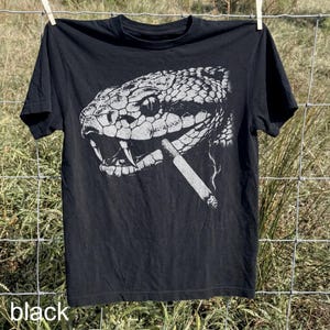 May include: Black t-shirt featuring a detailed, white graphic of a snake's head with a lit cigarette in its mouth. The snake's scales are intricately depicted. The word "black" is printed at the bottom of the shirt.