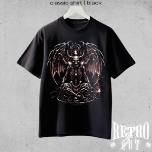 May include: Black t-shirt with a graphic print of a skeletal figure with wings sitting in a meditative pose. The figure has horns and glowing eyes. The graphic is in shades of gray and pink.