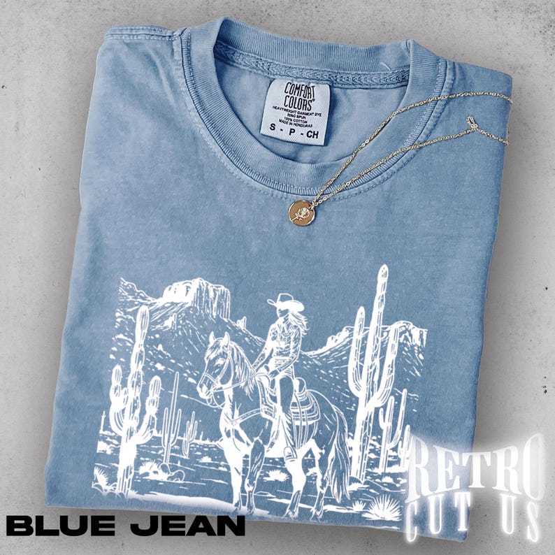 May include: A blue jean colored t-shirt with a white graphic of a cowgirl riding a horse through a desert landscape. The shirt has a Comfort Colors tag and a gold necklace with a pendant. The text "BLUE JEAN" is at the bottom.