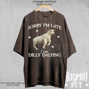 May include: A brown t-shirt with a vintage washed look. The shirt features a white sheep with the text "Sorry I'm Late I was Dilly Dallying" printed on it.