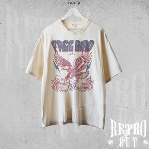 May include: A cream colored t-shirt with a vintage style graphic of a red and blue eagle with the text "FREE BIRD 1973" printed on the front.