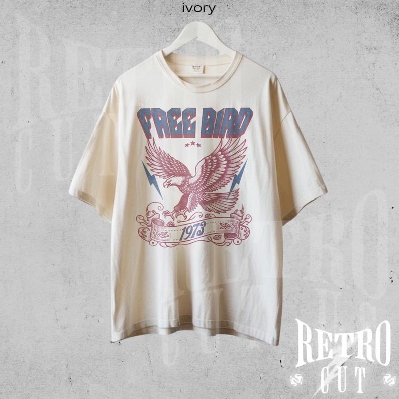 May include: A cream colored t-shirt with a vintage style graphic of a red and blue eagle with the text "FREE BIRD 1973" printed on the front.