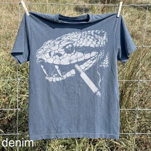 May include: A denim blue t-shirt featuring a detailed white graphic of a snake's head with a cigarette in its mouth. The word "denim" is printed at the bottom of the shirt. The shirt is hanging on a clothesline.