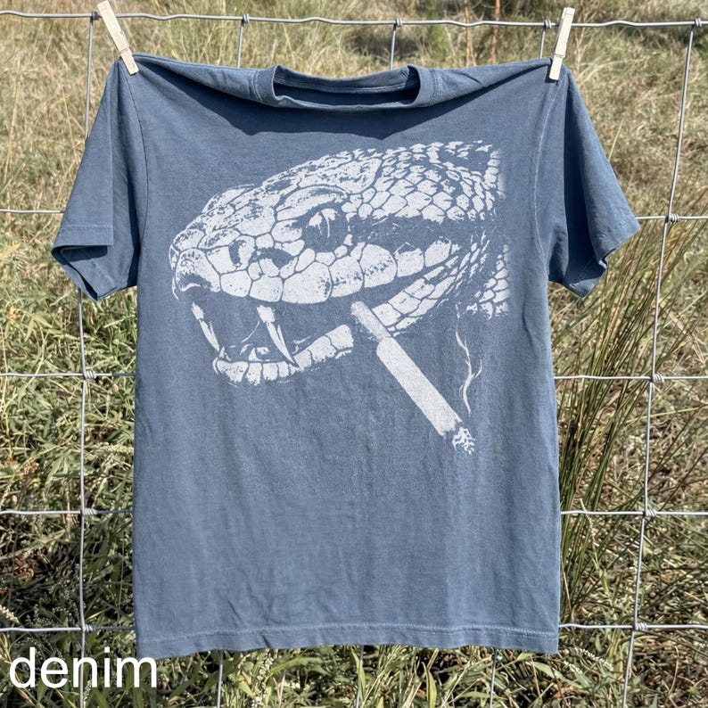 May include: A denim blue t-shirt featuring a detailed white graphic of a snake's head with a cigarette in its mouth. The word "denim" is printed at the bottom of the shirt. The shirt is hanging on a clothesline.