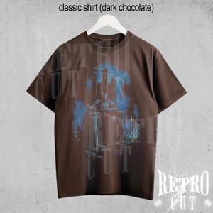 May include: A dark chocolate brown t-shirt with a blue and white graphic print of a castle and trees. The shirt has a crew neck and short sleeves. The text "Retro Cut" is printed on the bottom right corner of the shirt.