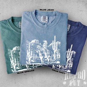 May include: Three folded t-shirts in blue spruce, blue jean, and true navy. Each shirt features a white graphic of a person on horseback in a desert landscape with cacti. The text "BLUE JEAN", "BLUE SPRUCE", and "TRUE NAVY" are visible.