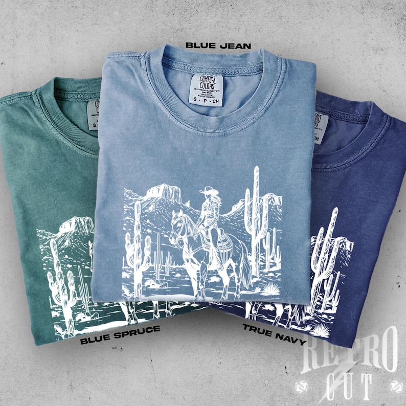 May include: Three folded t-shirts in blue spruce, blue jean, and true navy. Each shirt features a white graphic of a person on horseback in a desert landscape with cacti. The text "BLUE JEAN", "BLUE SPRUCE", and "TRUE NAVY" are visible.
