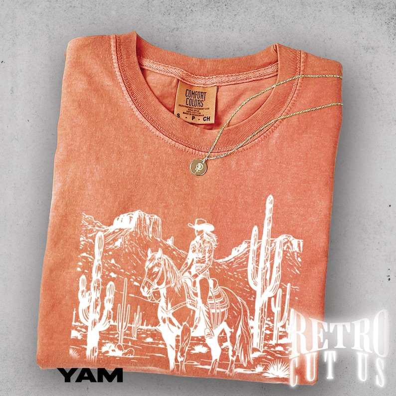 May include: An orange Comfort Colors t-shirt with a white graphic of a cowgirl on a horse in a desert landscape. A gold necklace with a pendant rests on the shirt. The text "YAM" is at the bottom left.