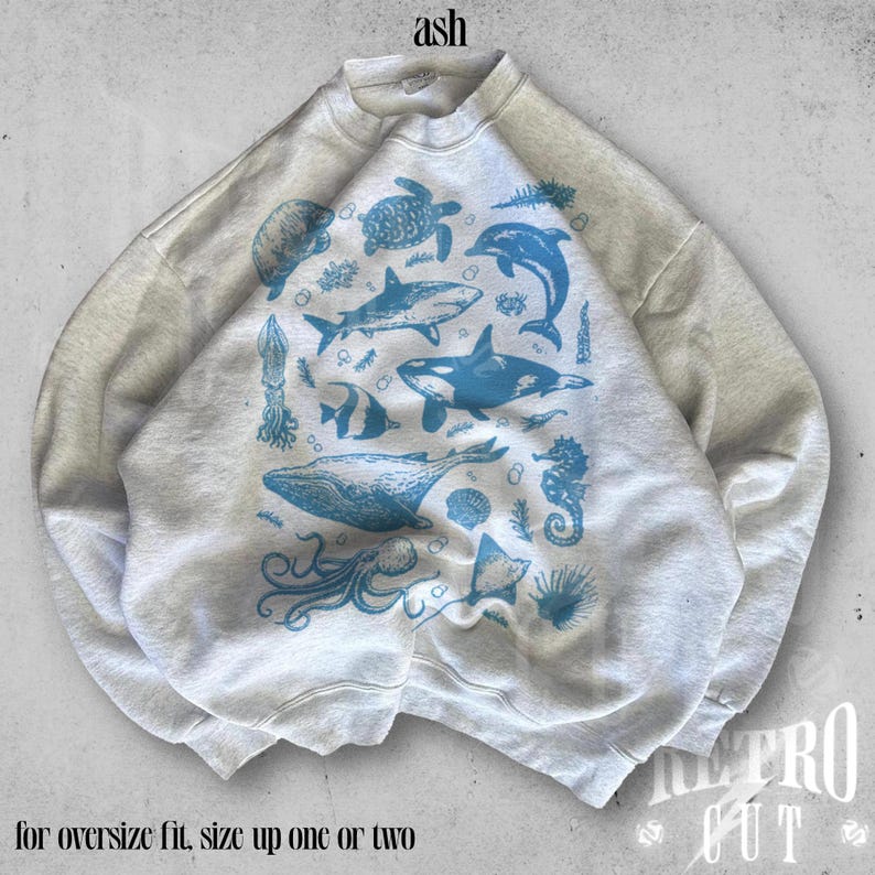 May include: A gray sweatshirt with a blue and white print of various sea creatures, including a whale, a dolphin, a shark, and a jellyfish. The text "ash" is printed on the top of the sweatshirt. The text "for oversize fit, size up one or two" is printed below the image.