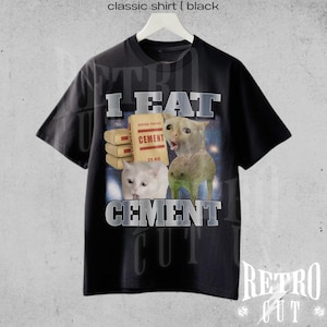Op de afbeelding: Zwart klassiek T-shirt met een afbeelding met de tekst "I EAT CEMENT" in zilveren letters. De afbeelding bevat een witte kat, een beige katachtig wezen en een zak cement. Het shirt heeft ook de tekst "Retro Cut".