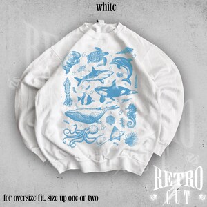 May include: White sweatshirt with a blue graphic print of various sea creatures, including a whale, shark, octopus, and seahorse. The text "for oversize fit, size up one or two" is printed below the graphic.