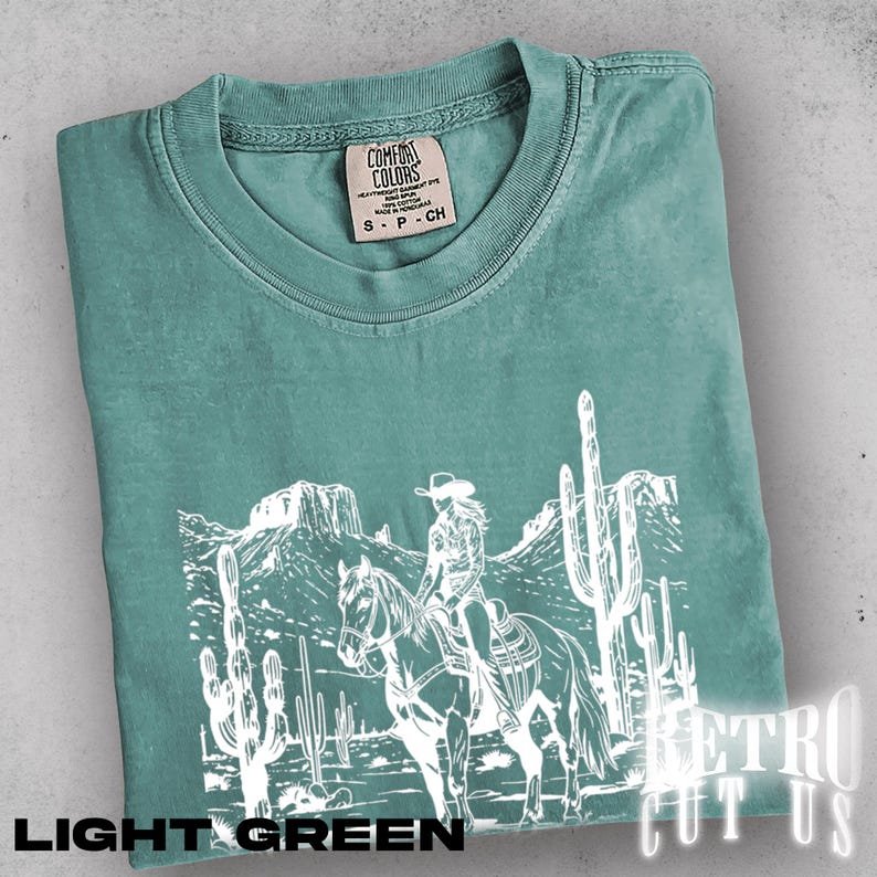 May include: Light green t-shirt with a white graphic of a person riding a horse through a desert landscape with cacti and mountains. The shirt has a crew neck and short sleeves. The text "LIGHT GREEN" is at the bottom.