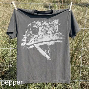 May include: Dark grey t-shirt featuring a large white graphic of a frog smoking a cigarette. The shirt is hanging on a clothesline. The word "pepper" is printed at the bottom of the shirt.