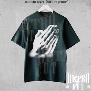 May include: Forest green classic t-shirt featuring a white graphic of praying hands. The shirt has short sleeves and a crew neck. The text "Retro Cut" is visible at the bottom right corner of the image. The shirt is hanging on a white hanger.