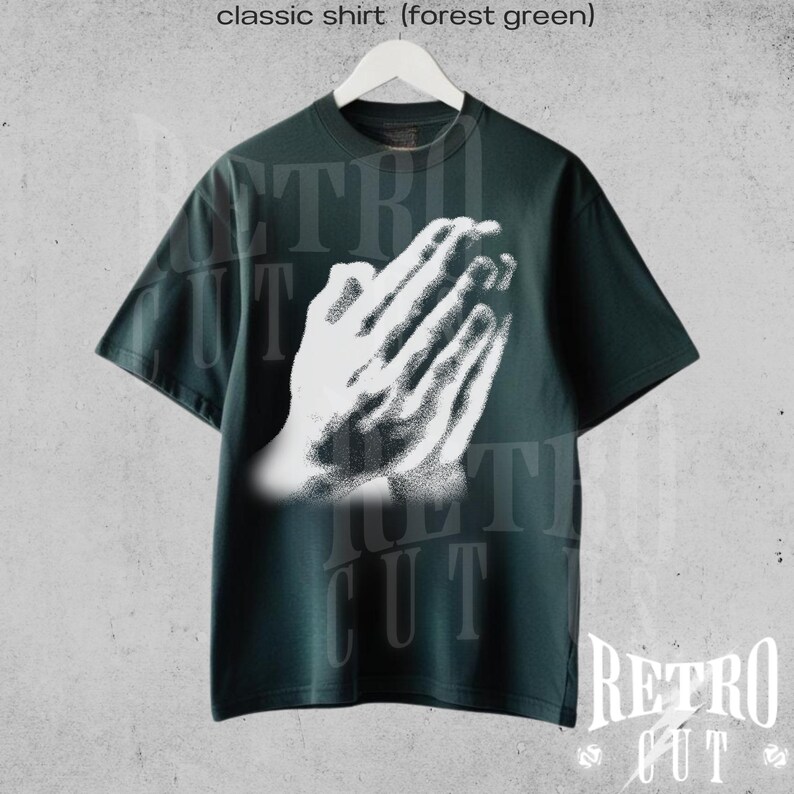 May include: Forest green classic t-shirt featuring a white graphic of praying hands. The shirt has short sleeves and a crew neck. The text "Retro Cut" is visible at the bottom right corner of the image. The shirt is hanging on a white hanger.