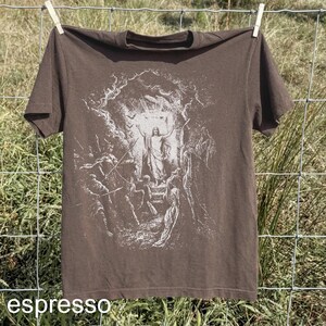 May include: Espresso-colored t-shirt with a white graphic depicting a religious scene. The image shows a figure with arms raised, surrounded by other figures. The shirt is hanging on a clothesline.