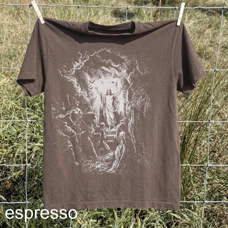 May include: Espresso-colored t-shirt with a white graphic depicting a religious scene. The image shows a figure with arms raised, surrounded by other figures. The shirt is hanging on a clothesline.