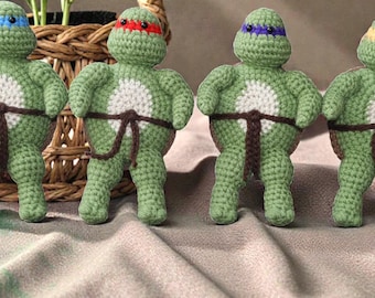 May include: Four handmade crochet figures of Teenage Mutant Ninja Turtles. Each turtle is green with a different colored mask: blue, red, purple, and yellow. They have white bellies and brown belts. The figures are standing on a gray surface.