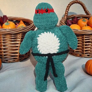 May include: A plush toy shaped like a turtle character with a dark green textured body, a red eye mask, a white circular patch on the chest, and a black belt tied around the waist. The toy has small black eyes and is made of soft yarn.