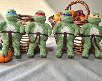 May include: Four handmade crochet dolls shaped like turtles, each featuring a light green body, a white belly patch, and a brown belt. The dolls wear masks in blue, red, purple, and orange. They are positioned in front of a woven wicker basket.