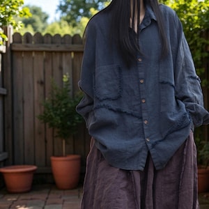 Puede incluir: Una camisa de lino azul oscuro con bordes deshilachados y dos bolsillos delanteros. La camisa está abotonada y combinada con una falda larga y fluida de color púrpura oscuro. El atuendo se usa al aire libre, con una valla de madera y plantas en macetas en el fondo.